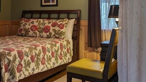 A bedroom with a wooden ceiling, a bed with floral bedding, a chair, a trash can, and a window with curtains. - Cabin surrounded by Lustrous Pine trees_Cabin 14 (Bar Harbor)