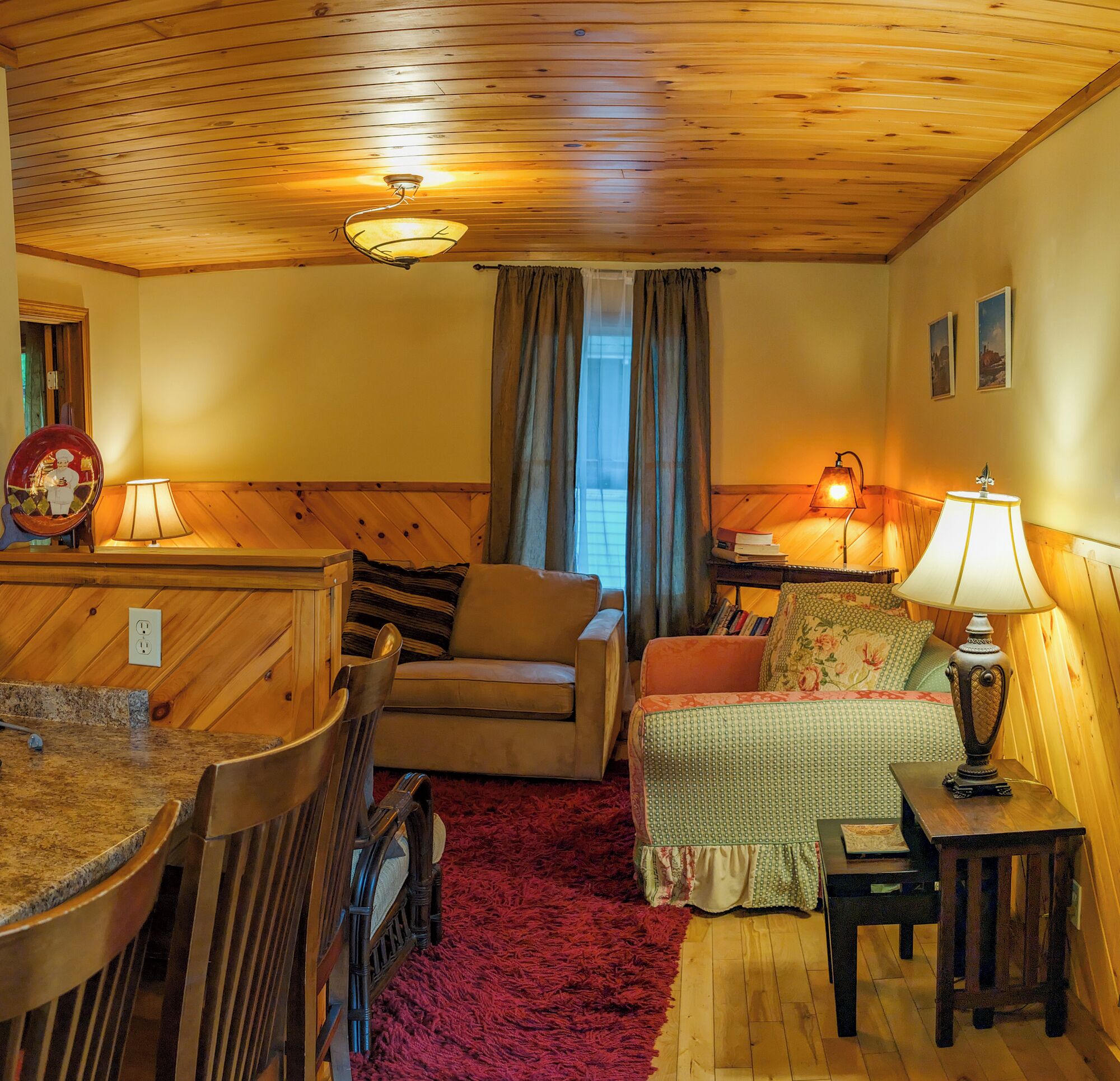 A cozy cabin interior with a wooden ceiling, a dining table with chairs, a sofa, a small table with a lamp, and a window with curtains.