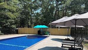Une piscine entourée d’une clôture en bois, avec une terrasse en bois et des espaces de détente en plein air agrémentés de parasols.
