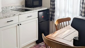 A compact kitchen with white cabinets, a black refrigerator, a microwave, and a coffee maker on the countertop. There's a wooden dining table with a chair, and a window with sheer curtains and a checkered valance.