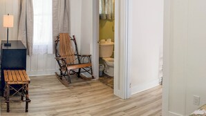 A room with a wooden floor, a rocking chair, a small table, a lamp, and a door leading to a bathroom with a toilet and a sink.