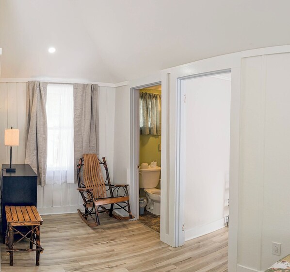 A room with a wooden floor, a rocking chair, a small table, a lamp, and a door leading to a bathroom with a toilet and a sink. - New & Shiny_Dream Holiday_Cabin 19 (Bar Harbor)