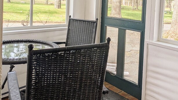 A covered porch with two wicker chairs and a small table, overlooking a grassy area with trees. - New Cabin to ENJOY!_Cabin 20 (Bar Harbor)