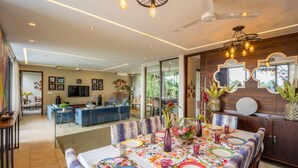 A modern dining area with a large table set for a meal, a kitchen in the background, and a living area with a sofa and TV. - Avior by Hireavilla - 4BR w Infinity Pool in Parra (Parra)