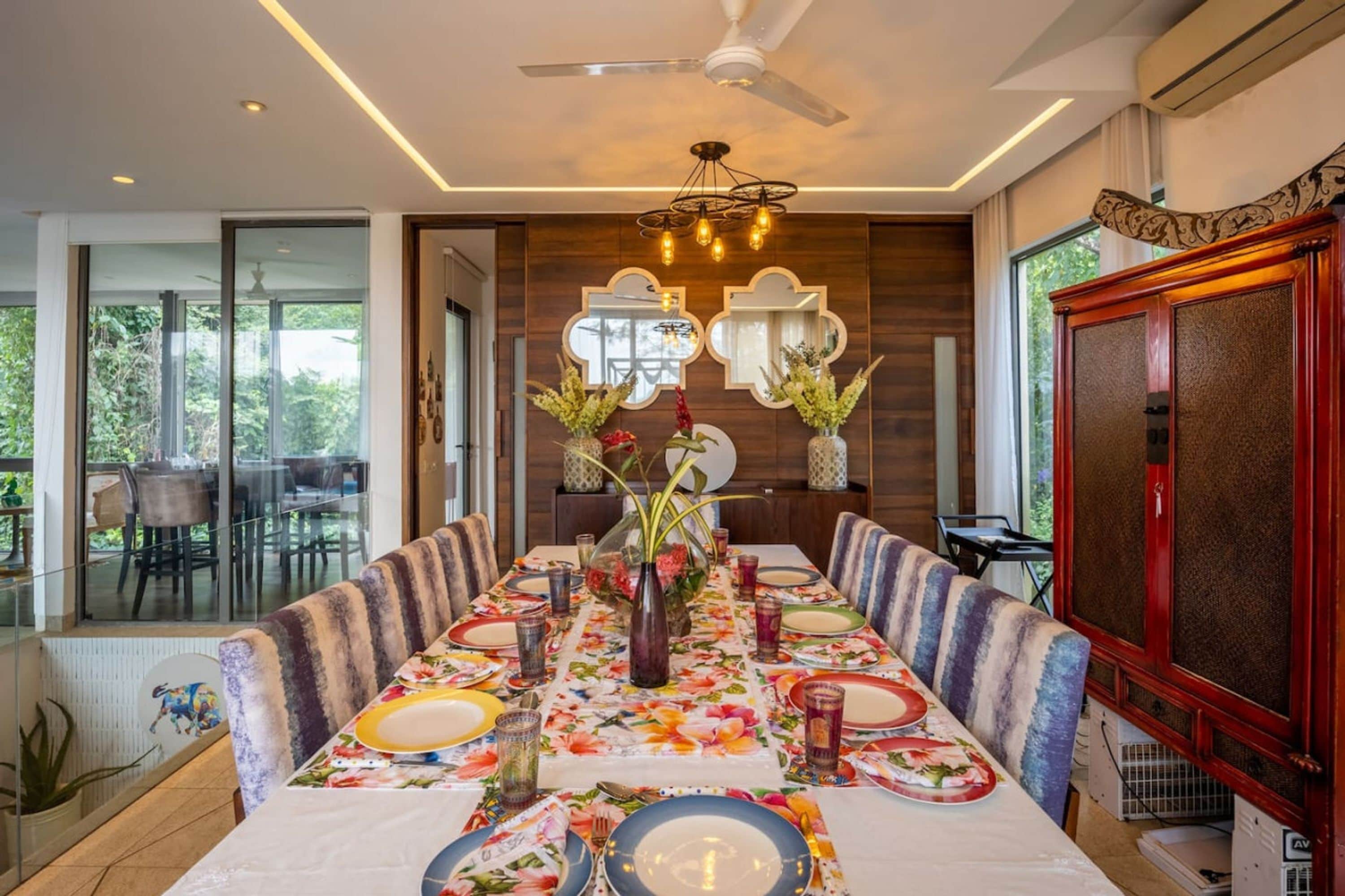 A dining area with a long table set for a meal, surrounded by chairs, with a ceiling fan and decorative lighting above.