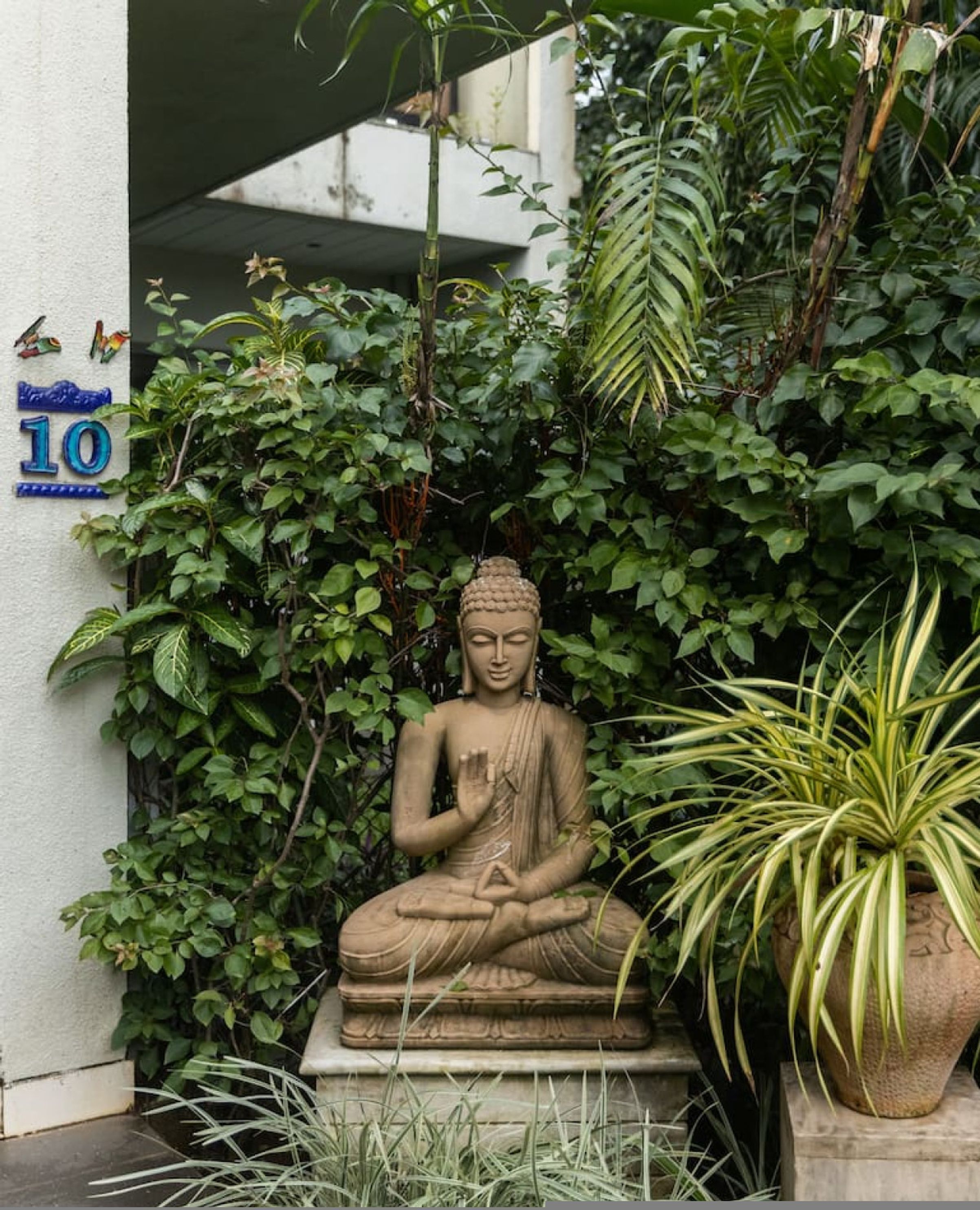 A Buddha statue in a garden setting with lush greenery and a building in the background.