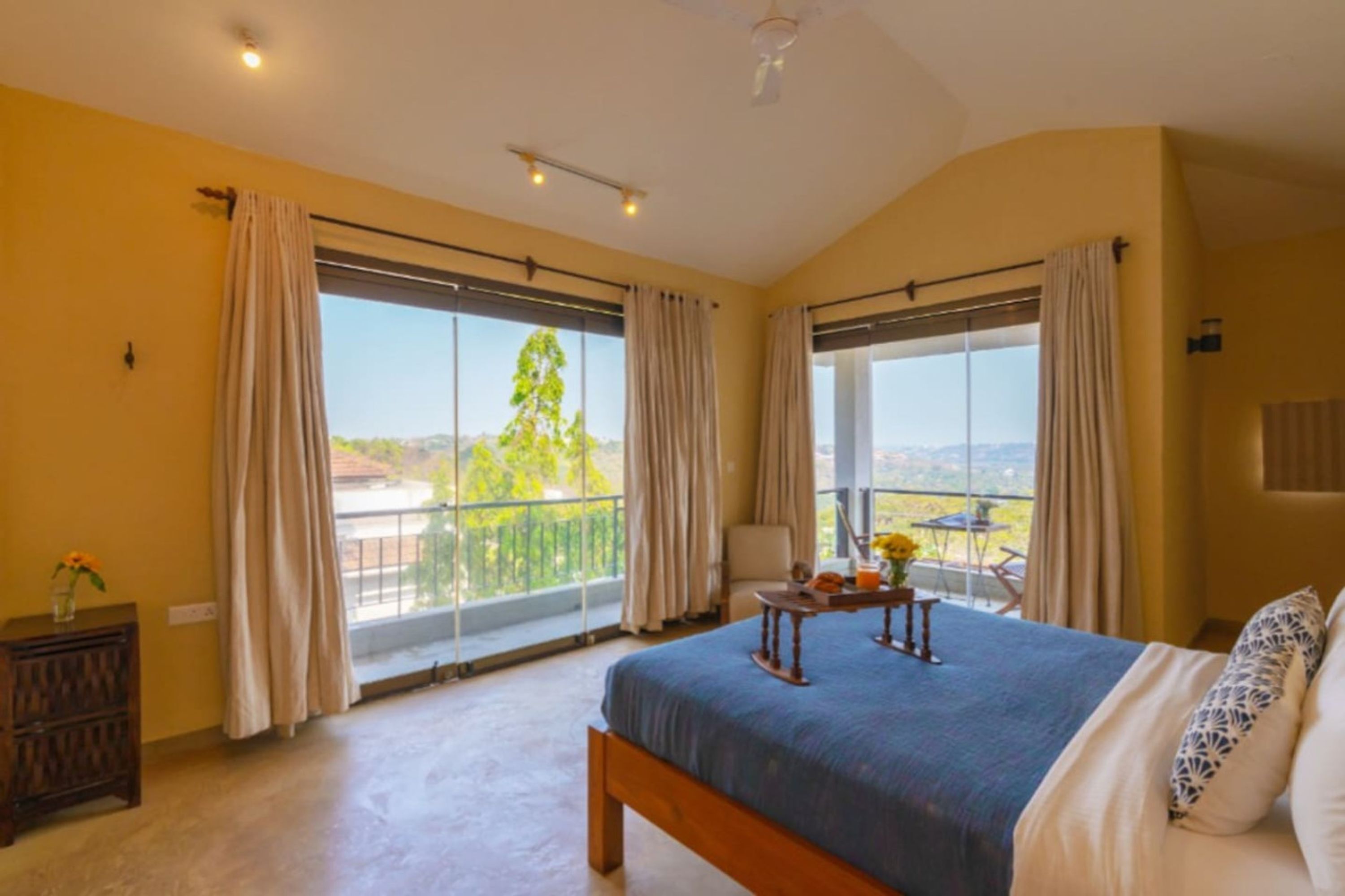 A bedroom with a large bed, two windows with curtains, a small table with a vase, and a view of greenery outside.