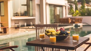 A poolside table with a bowl of fruit and glasses of orange juice, a wooden chair, and a modern building in the background. - Cliffhouse by Hireavilla -5br w Pvt Pool in Saipem (Candolim)