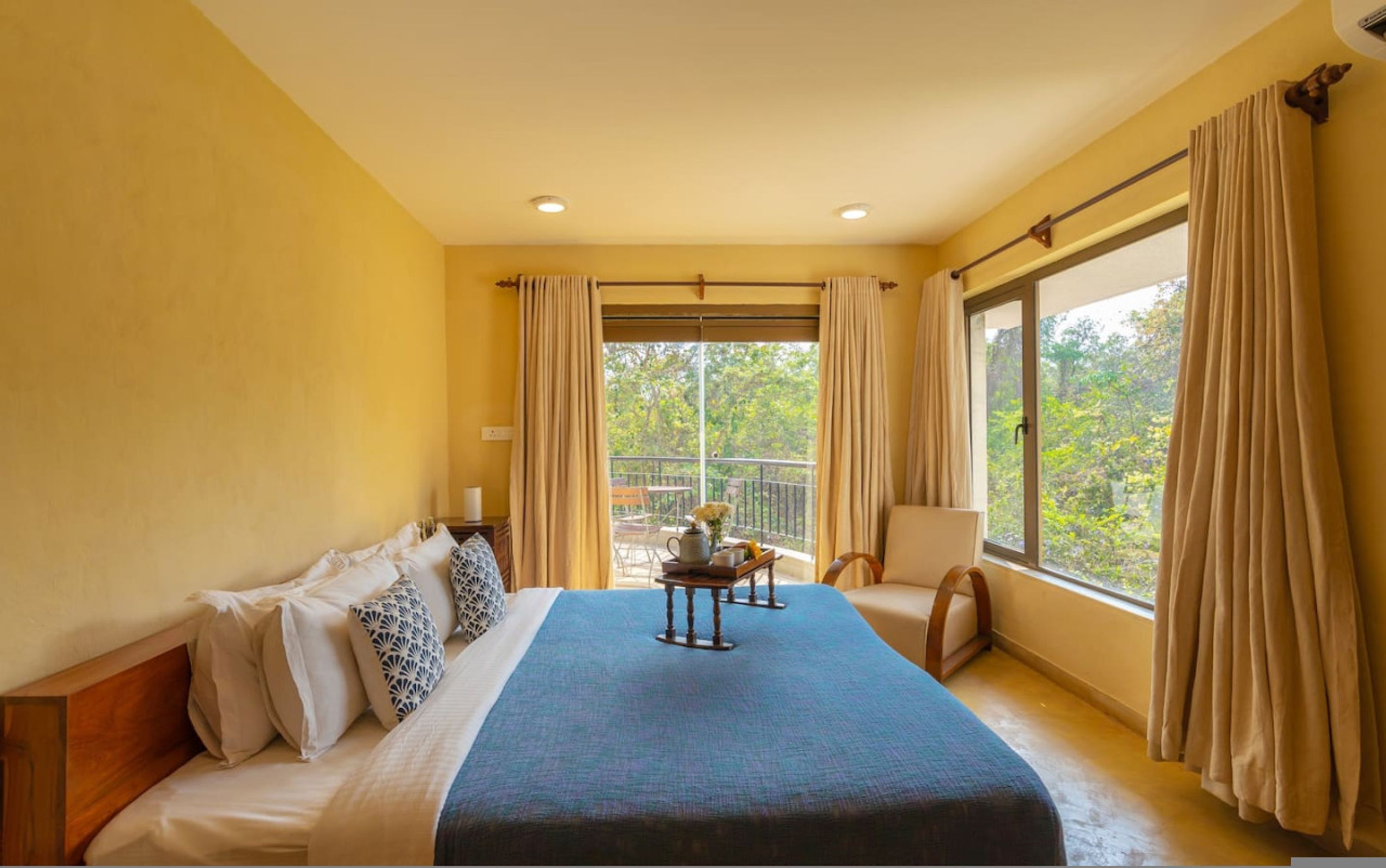 Una habitación de hotel con una cama grande, una mesita con un florero y flores, una silla y un balcón con cortinas.