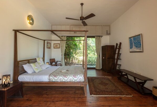 A four-poster bed with a wooden frame and a canopy, a ceiling fan, a wooden bench, and a view of greenery through the window.