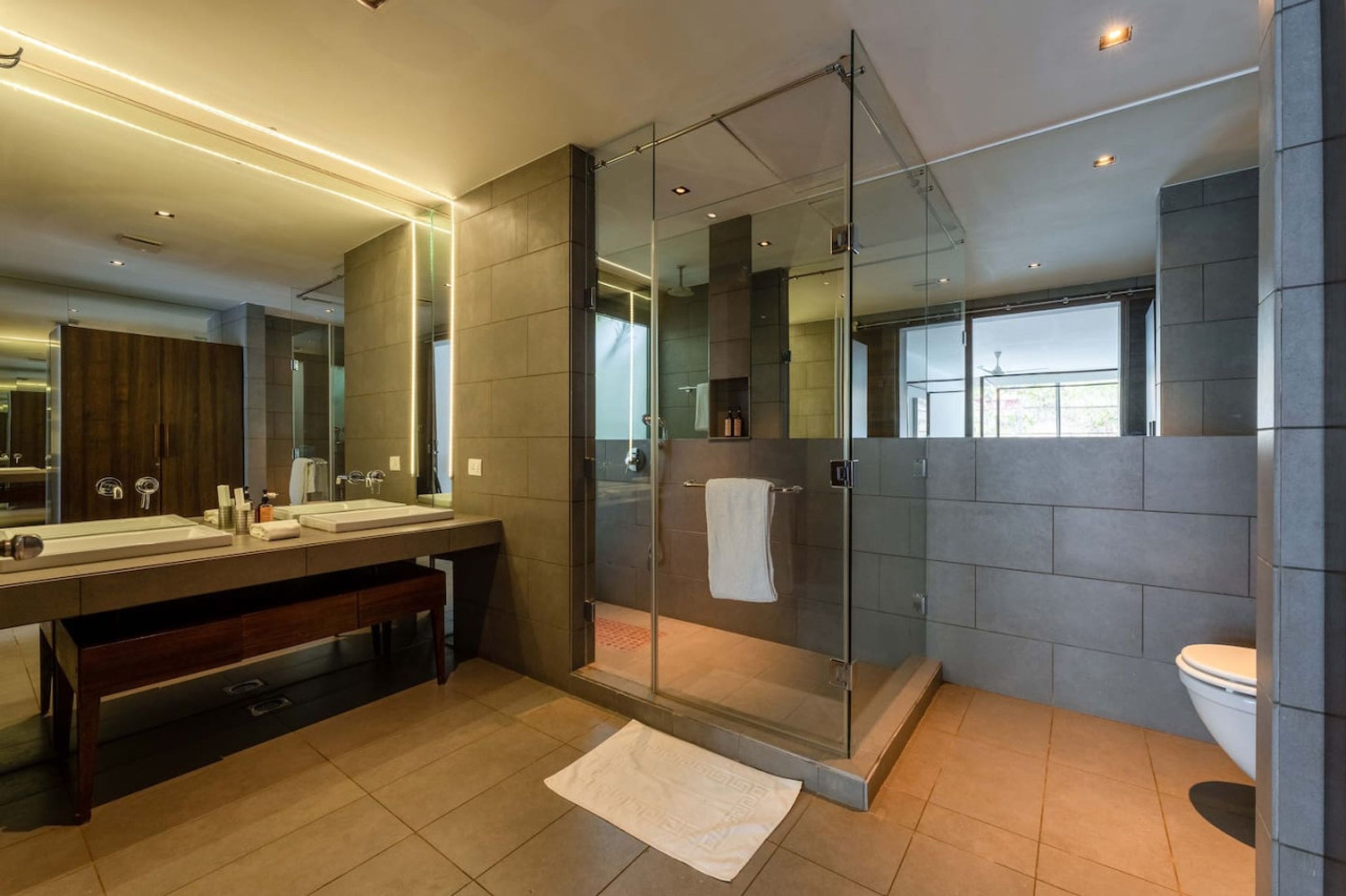 A modern bathroom with a glass-enclosed shower, a large mirror, and a vanity with a bench.