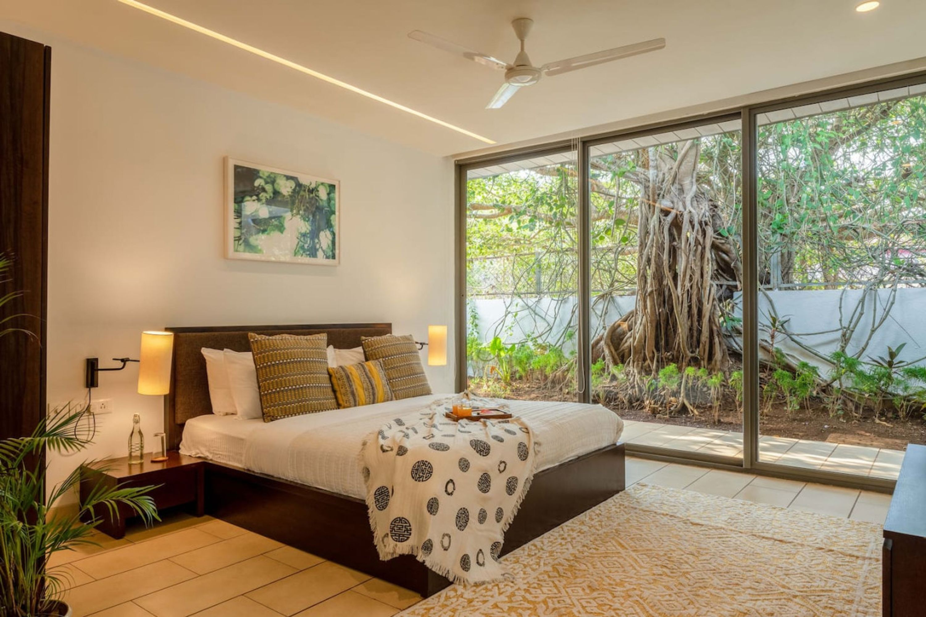 A modern bedroom with a large bed, a ceiling fan, and a view of a garden through large windows.