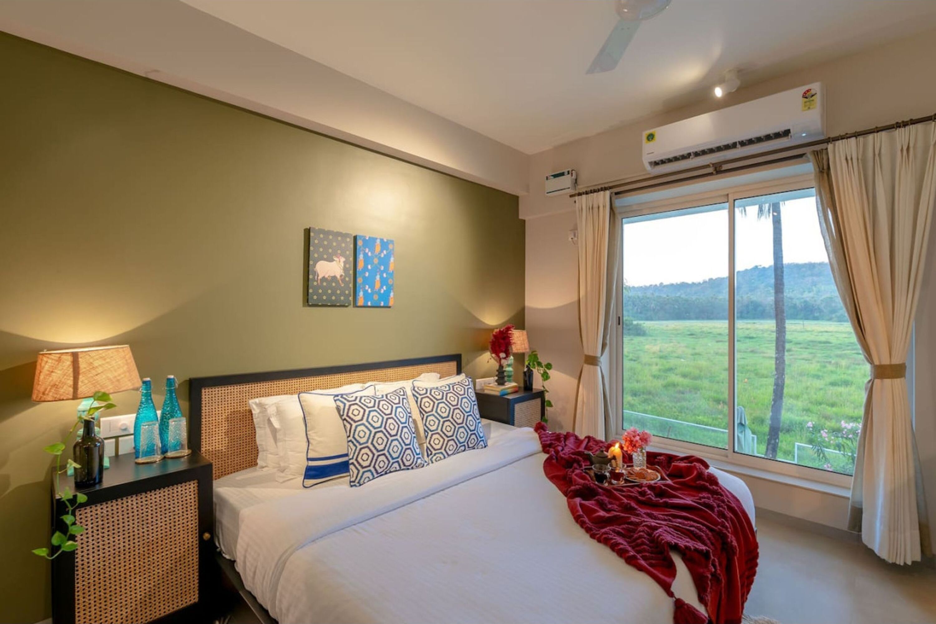 Un dormitorio con una cama grande, una mesita de noche con lámpara, un aire acondicionado instalado en la pared y una ventana con vistas a un paisaje verde.