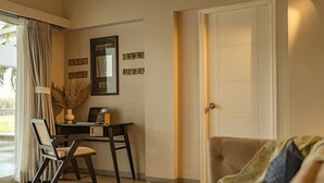 A living room with a sofa, a desk with a chair, a door, and a window with curtains. - Legacy by Hireavilla Natgeo Spot w Field Views (Siolim)