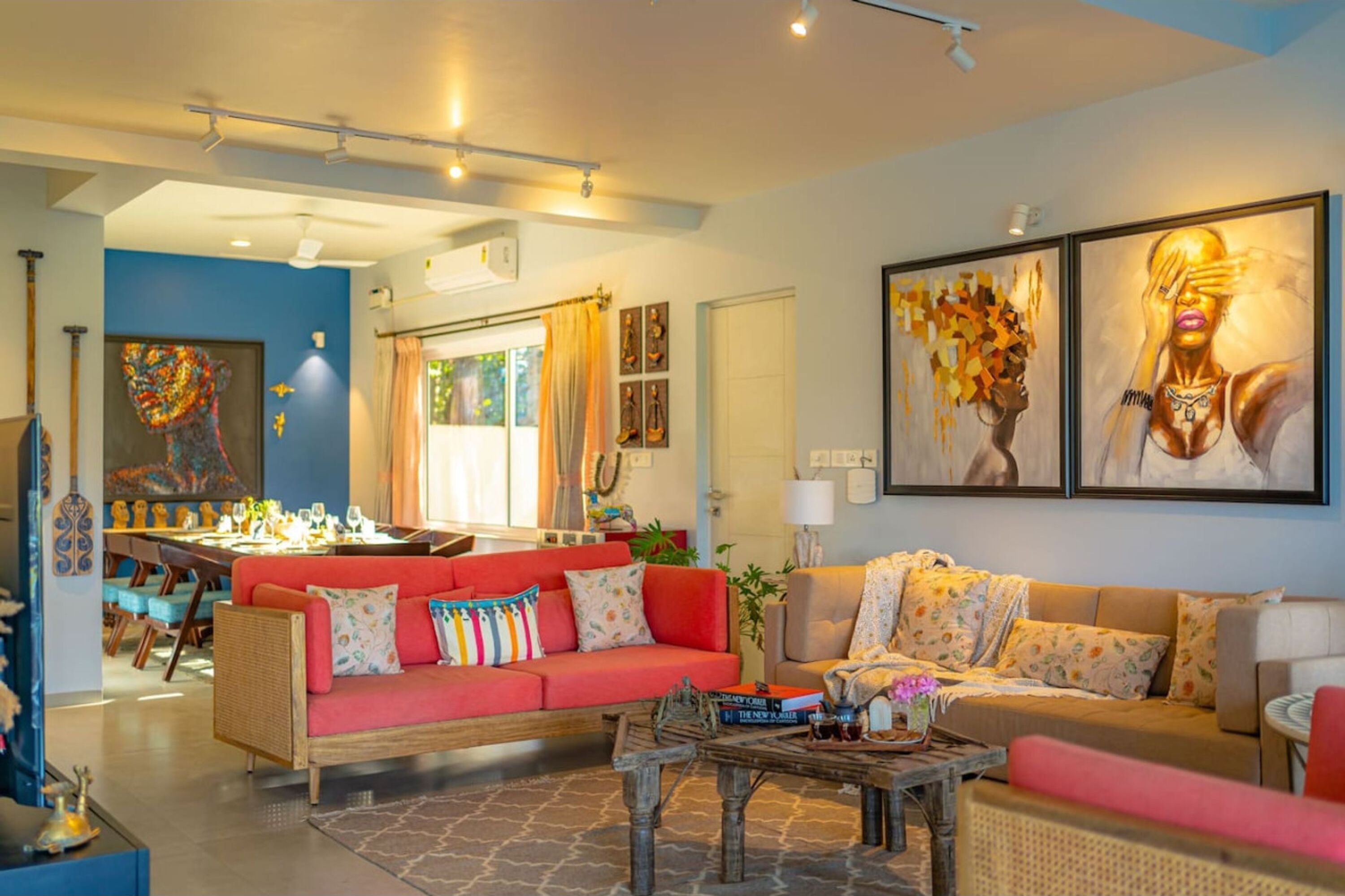 Un vaste salon comprenant un canapé rouge, un canapé beige et une table basse. Deux grandes peintures ornent le mur. On y trouve également un coin repas avec une table et des chaises en bois, et une cuisine visible au fond.