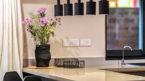 Une cuisine moderne avec un îlot central, des tabourets de bar noirs, des luminaires suspendus et un vase rempli de fleurs.