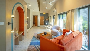 Un salon moderne avec un canapé, un fauteuil et une table basse. On y trouve une grande fenêtre avec des rideaux, un ventilateur de plafond et un climatiseur fixé au mur.