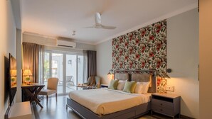 Una habitación de hotel moderna con una cama grande, un escritorio con una silla, una mesita pequeña, un ventilador de techo y un balcón con cortinas.