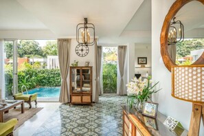 A spacious living area with a large window, a pool, a wooden cabinet, and a round mirror. - Sol by Hireavilla - 2BR w Private Pool in Anjuna (Vagator)