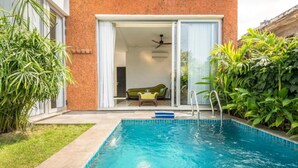 Ein Schwimmbecken mit klarem, blauem Wasser, umgeben von einer Ziegelwand und einer Glastür, die zu einem Raum mit einem grünen Sofa führt.