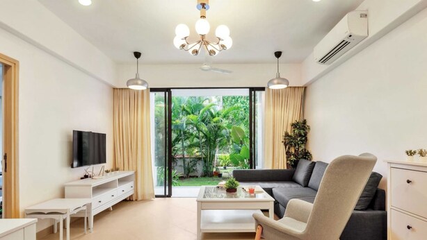 A modern living room with a sofa, a coffee table, a TV, and a view of a garden through large windows. - Cozy 1BR With Pool - Only 1km From Candolim Beach (Candolim)