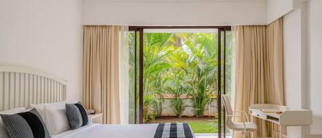 A modern hotel room with a large bed, a desk, and a view of a lush garden.