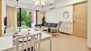 A modern living room with a dining area, a grey sofa, a white cabinet, and a large window with a view of greenery. - Cozy 1BR With Pool - Only 1km From Candolim Beach (Candolim)