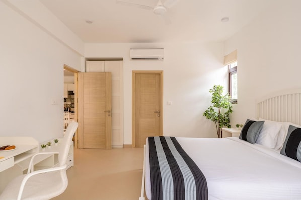 A modern bedroom with a large bed, a desk, and a chair. There is a wardrobe, a plant, and an air conditioning unit.