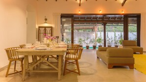 A dining area with a wooden table and chairs, a sofa, and a window with a view of plants and a pergola. - Salt by Hireavilla - 4BR w Heated Pool in Siolim (Siolim)