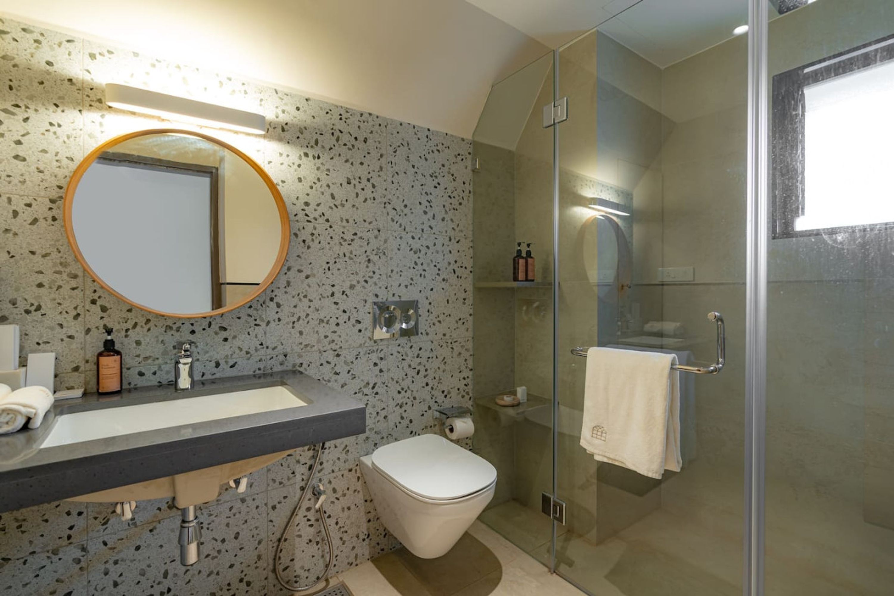 A modern bathroom with a granite countertop sink, a round mirror, a toilet, and a glass-enclosed shower area.
