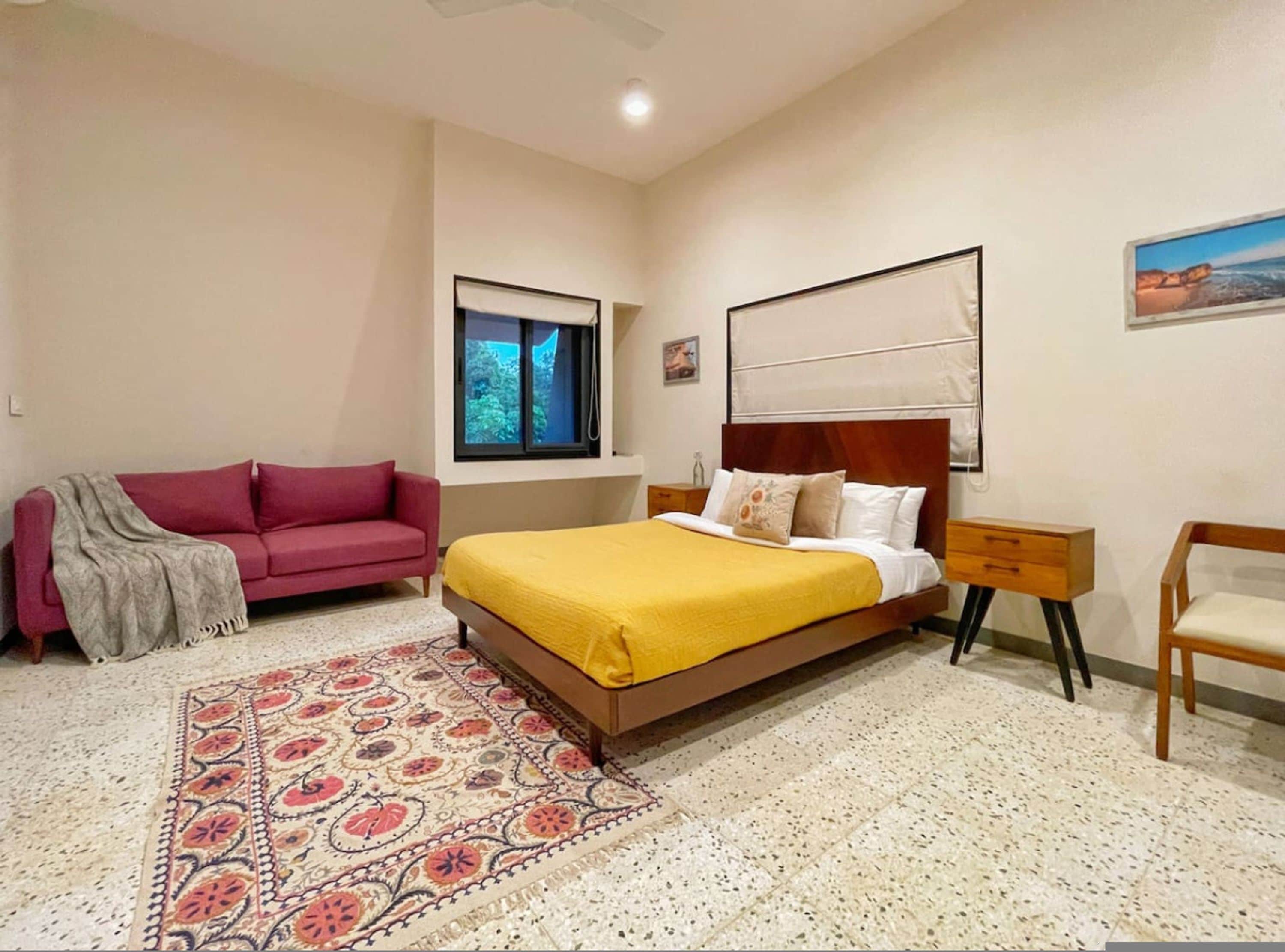 Un dormitorio con cama, un sofá rojo, una mesita de noche de madera, una silla, un tapete estampado, un ventanal y un cuadro en la pared.