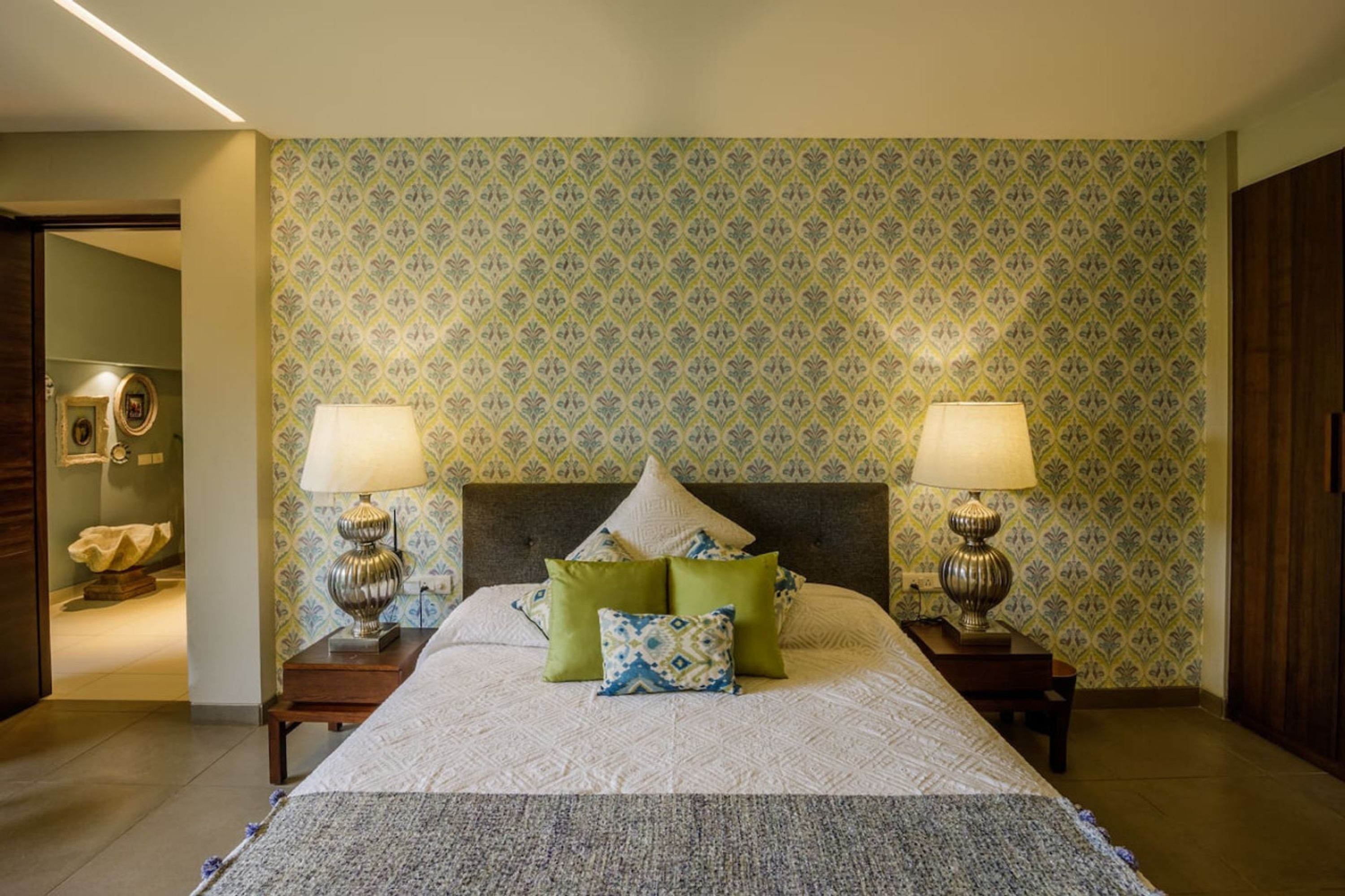 A neatly arranged bedroom with a large bed, two bedside tables with lamps, patterned wallpaper, and a wooden wardrobe.
