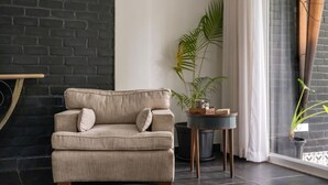 Una sala de estar con un sofá color beige, una mesita redonda, una planta en maceta y una ventana con cortinas.
