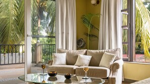 Una sala de estar con un sofá color beige, dos cojines blancos, una mesa de centro de vidrio con un tazón y copas de vino, y vista a palmeras a través de amplias ventanas.