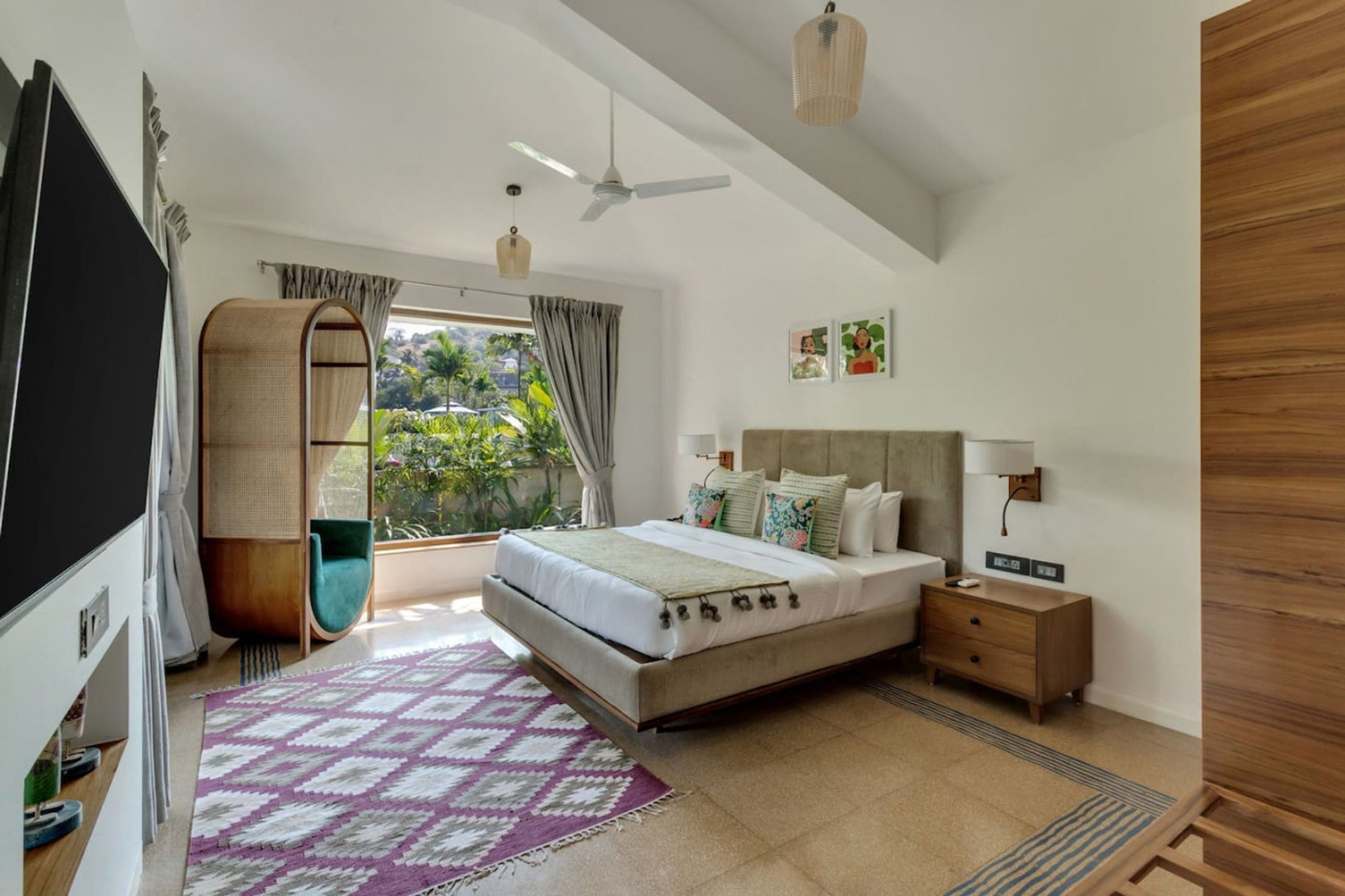 A modern bedroom with a large bed, a bedside table, a TV, and a view of greenery outside.