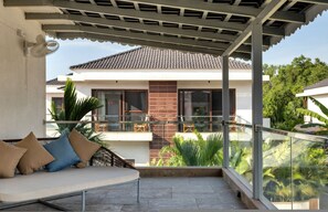 A covered balcony with a sofa, glass railing, and a view of a house with a tiled roof. - Inaari - 5bhk With Private Pool Near Anjuna Beach (Anjuna)