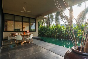 A modern indoor pool area with a dining table and chairs, surrounded by lush greenery and a clear pool.