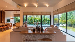 A modern living room with a large window, a ceiling fan, and a dining area. - Puravida I -luxury 5bhk w Pvt Pool in Anjuna (Anjuna)