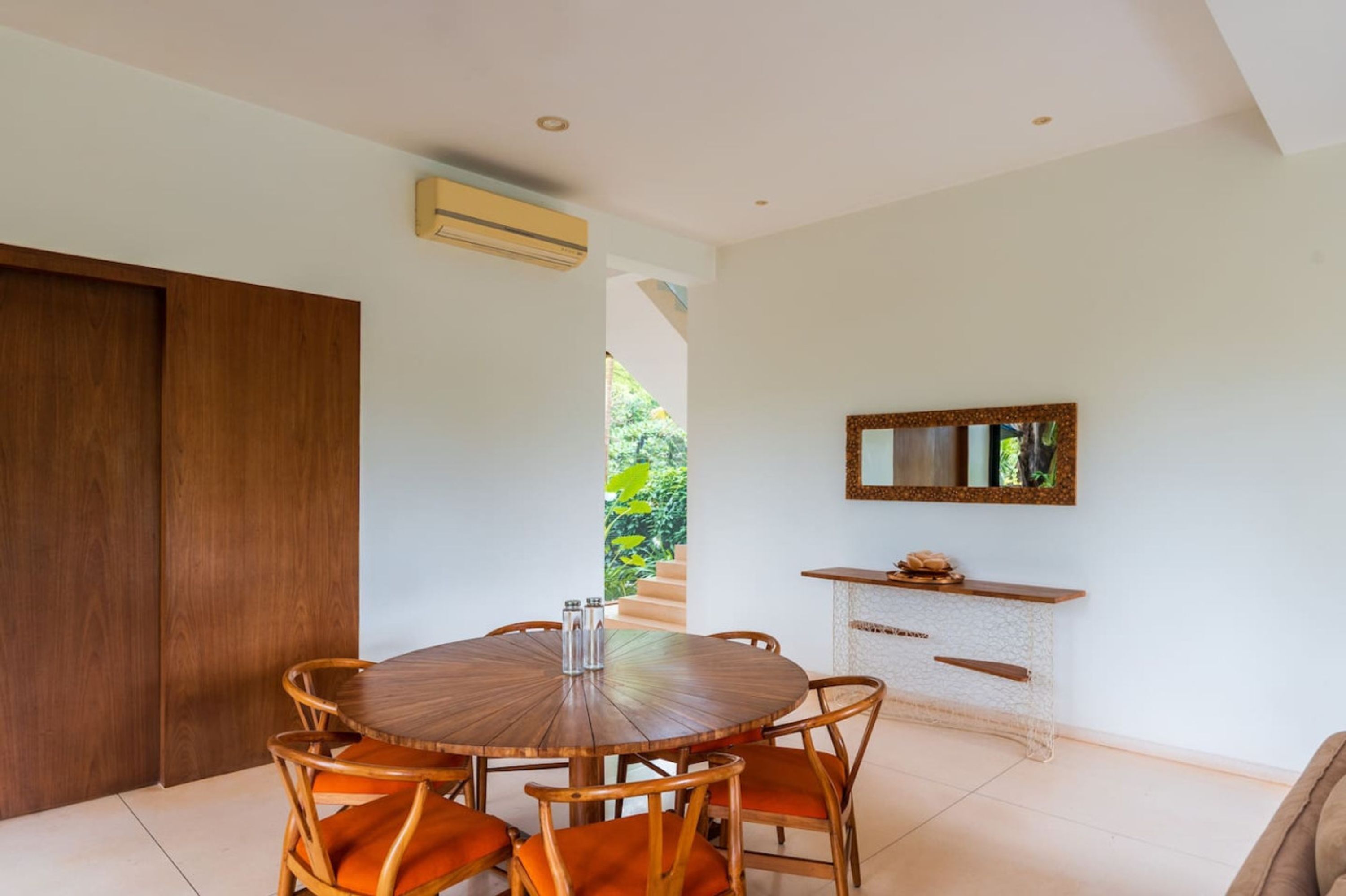 A dining area with a round wooden table and wicker chairs, a small console table with a mirror, and a view of a staircase leading to a garden.
