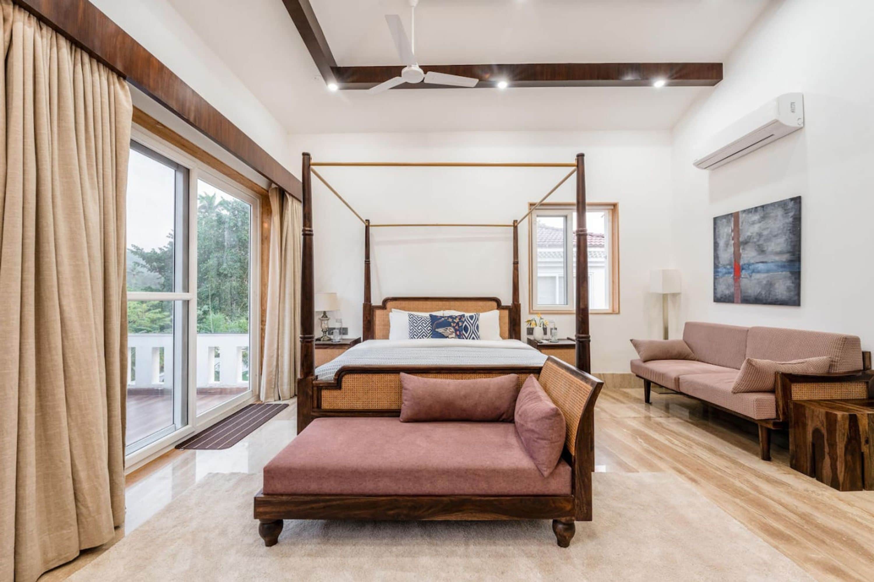 Ein Bett mit Baldachin und Holzrahmen, eine Couch und ein Couchtisch in einem hellen Raum mit großen Fenstern und Deckenventilator.