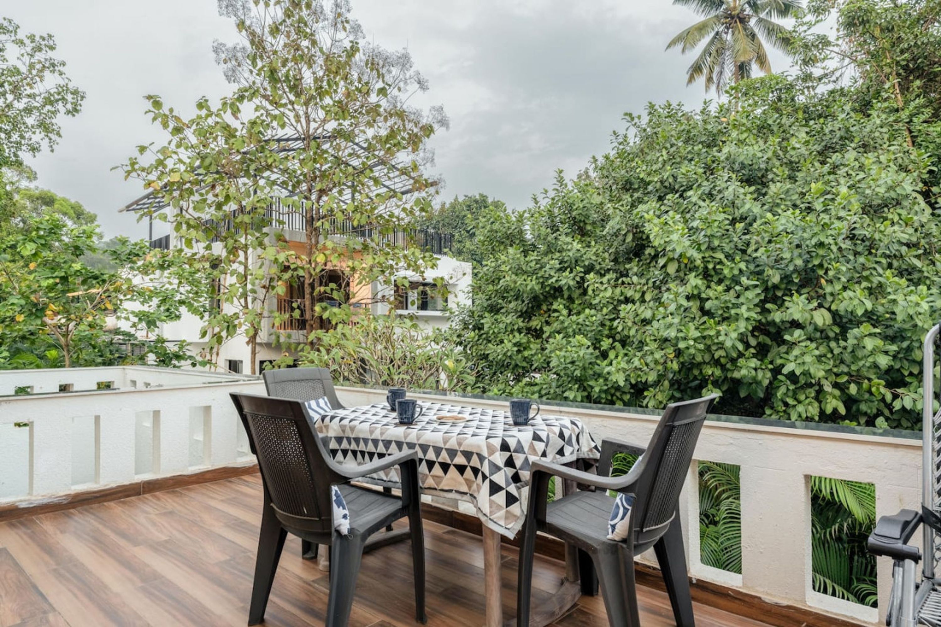 A covered outdoor seating area with a table and chairs, surrounded by lush greenery and a white building in the background.