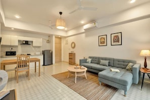 A modern living room with a grey sofa, a wooden dining table, and a kitchen in the background. - Sol Banyan by Hireavilla - 1BR w Pool in Candolim (Candolim)