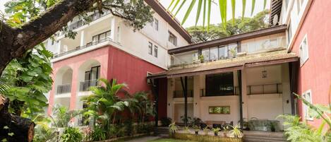 Un edificio con fachada roja, un patio cubierto con mesas al aire libre y un jardín cuidado con diversas plantas y un sendero.