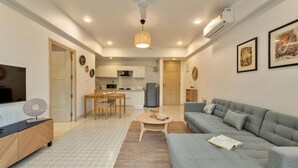 A modern living room with a grey sofa, a coffee table, and a TV on a wooden cabinet. There is a kitchen area with a dining table and chairs, and a ceiling fan. - Sol Banyan by Hireavilla - 1BR w Pool in Candolim (Candolim)