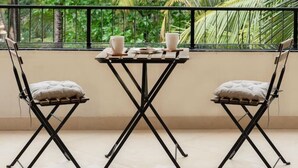 A balcony with two chairs and a table, overlooking a lush garden with palm trees. - Sol Banyan by Hireavilla - 1BR w Pool in Candolim (Candolim)