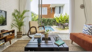 A living room with a TV, books, a coffee table, a sofa, a potted plant, and a view of a pool and building. - Nova by Hireavilla - 2BR w Private Pool in Anjuna (Vagator)