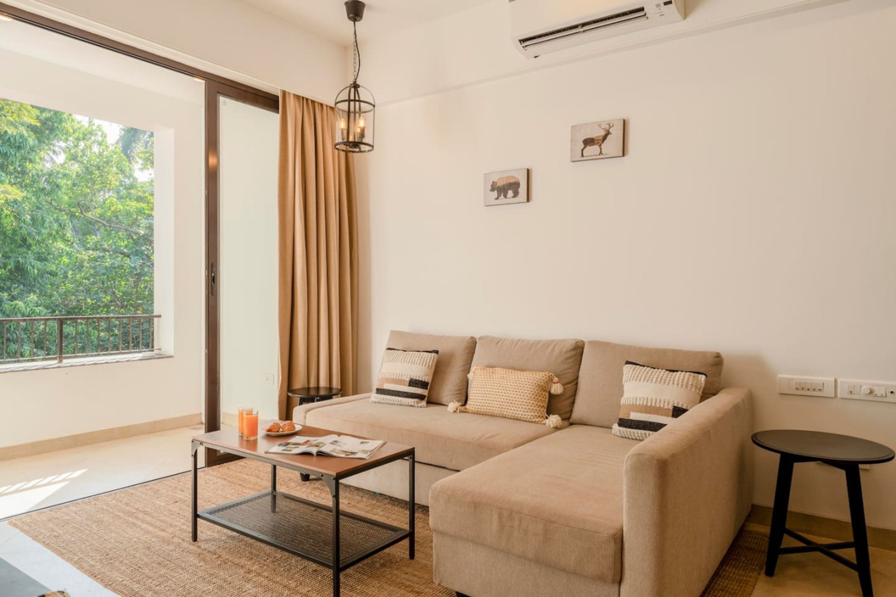 A modern living room with a sofa, a coffee table, and a small table. There is a window with curtains, a wall-mounted air conditioner, and a picture of a deer.