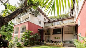 Un edificio con fachada roja, un patio cubierto con mesas al aire libre y un jardín cuidado con diversas plantas y un sendero.