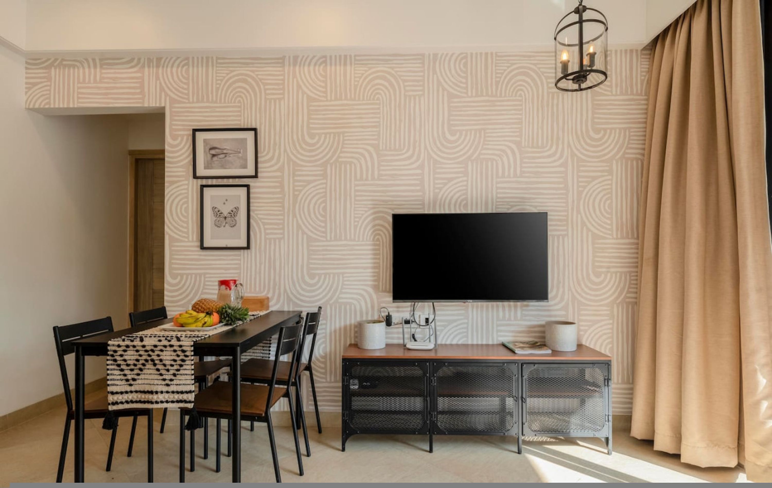 A modern living room with a flat-screen TV mounted on the wall, a dining table with chairs, and a small side table with a speaker and a remote control.