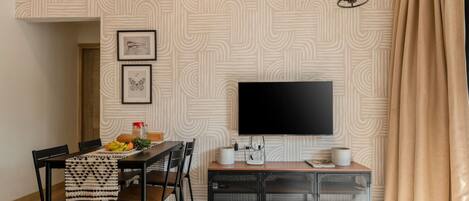 A modern living room with a flat-screen TV mounted on the wall, a dining table with chairs, and a small side table with a speaker and a remote control.